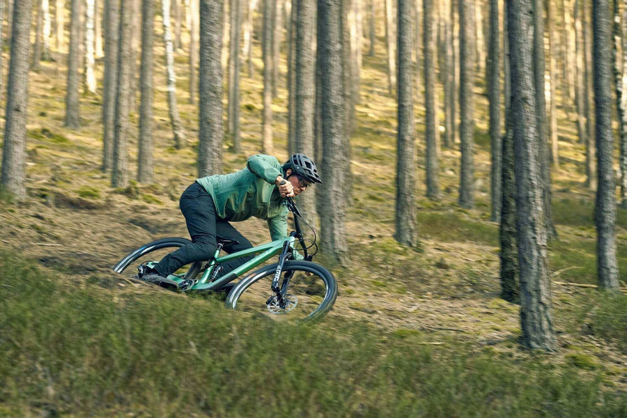 Ein Mann fährt mit dem BUEX Pro 2.0 Mountainbike in den Wald. Robuste MTB Hose aus Bio-Nylon auf Basis von Rizinus Öl. Dynamischer Schnitt, abriebfest, mit Taschen und reflektierenden Elementen. Ideal für Gravelbike, Mountainbike & Roadbike. Nachhaltige Radbekleidung von TRIPLE2.